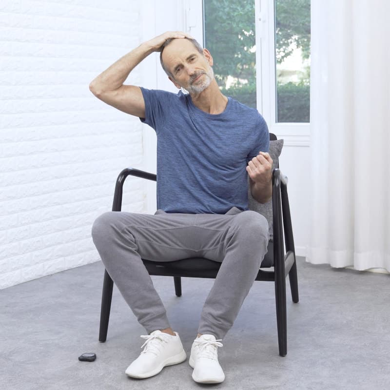Roland Liebscher-Bracht is sitting in a chair making a fist with his left hand and pulling his head to the right with his right hand.