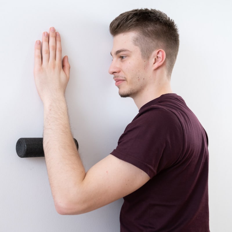 A man is foam rolling his forearm against a wall.