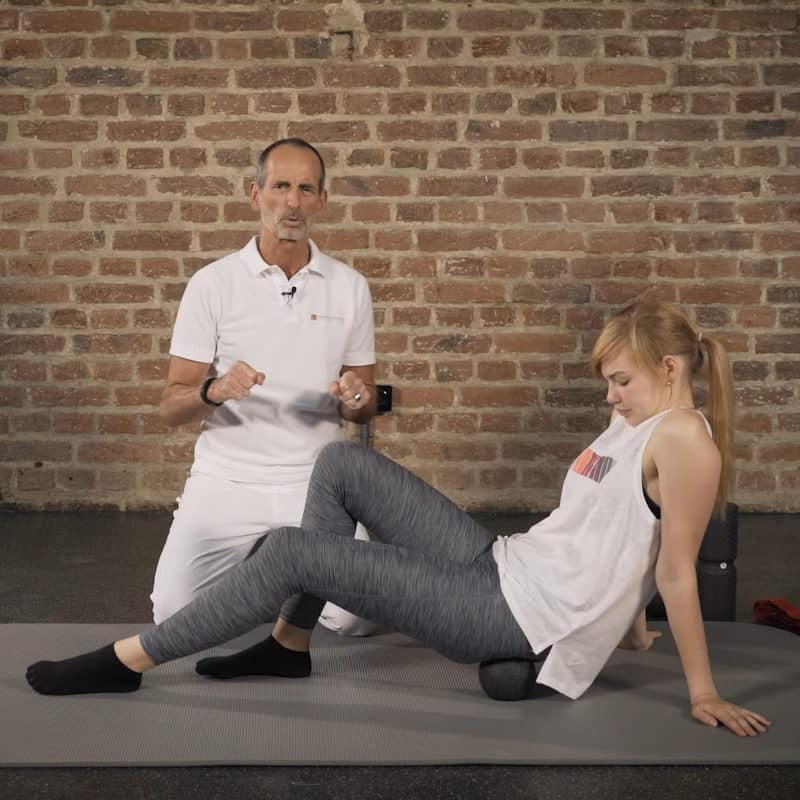 A girl is sitting on the Midi Massage Ball as Roland Liebscher-Bracht looks on.