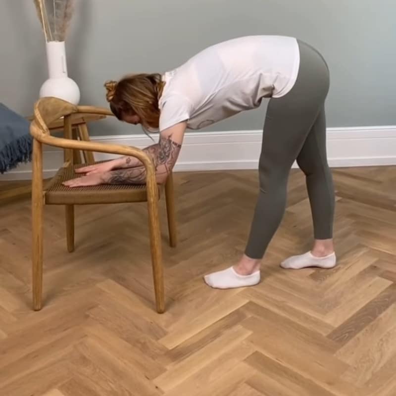 A girl is stretching her legs with her forearms on a chair.