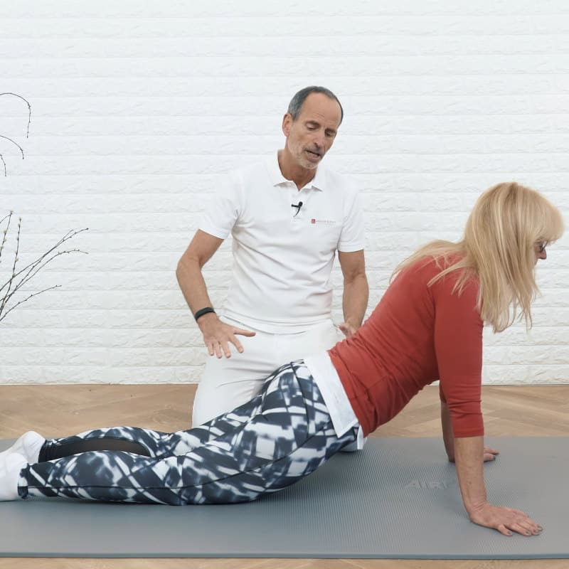 A woman is stretching in seal pose while Roland Liebscher-Bracht looks on.
