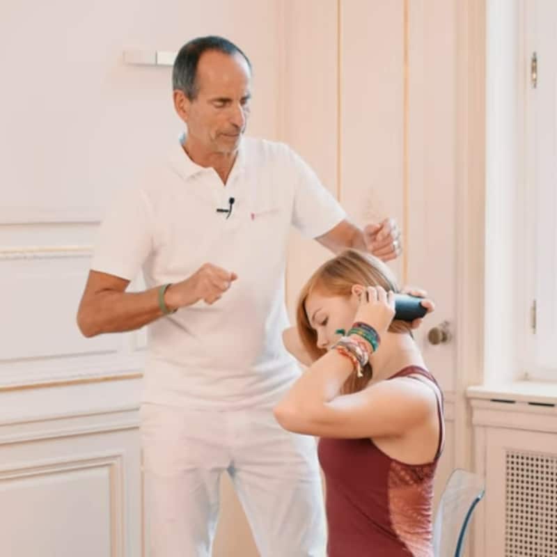 A woman is massaging the left side of her neck with the Mini Foam Roller as Roland Liebscher-Bracht looks on.