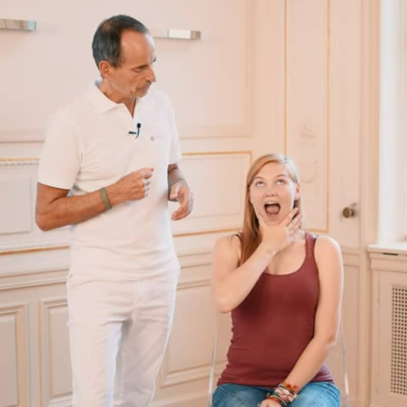 A woman is pulling her chin down with her thumb and index finger while Roland Liebscher-Bracht looks on.