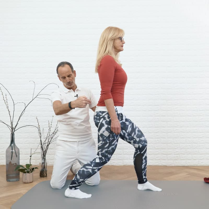 A woman is standing with her left leg in front of her bending at the knees while Roland Liebscher-Bracht looks on.