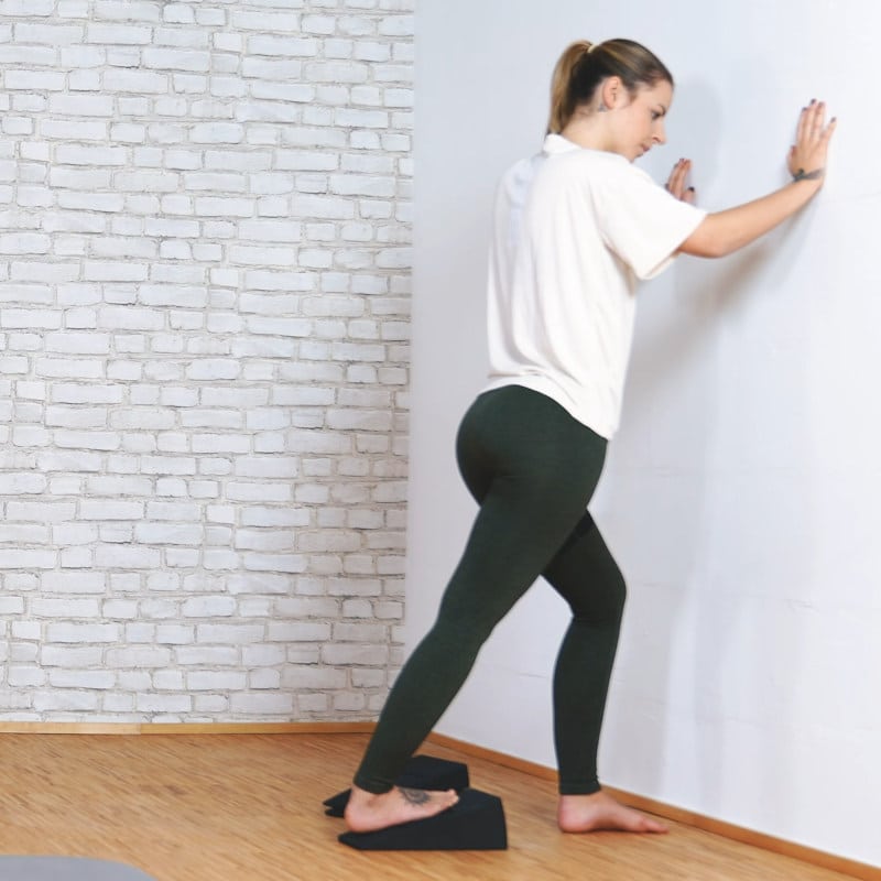 A girl is standing with her right leg on the Knee Hero and her hands against the wall.