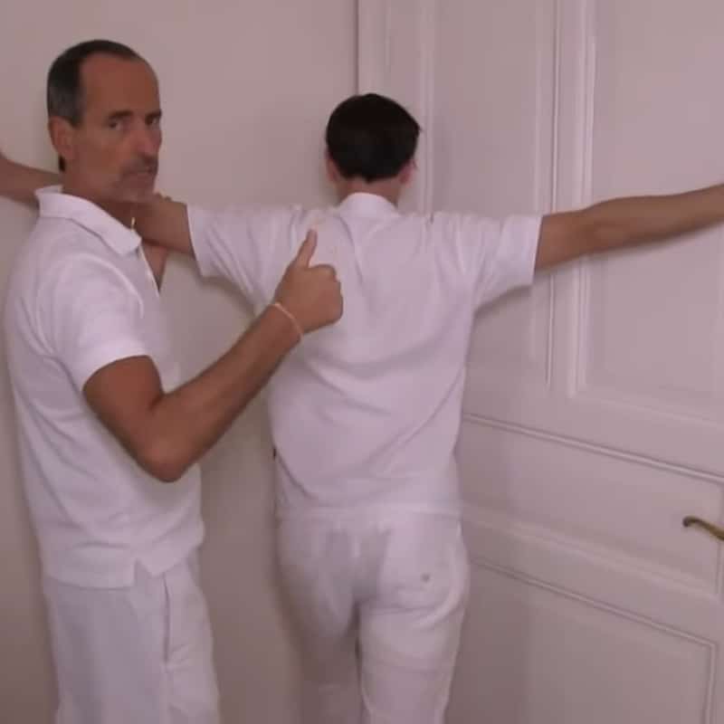 A man is standing in a corner with his hands on the wall while Roland Liebscher-Bracht looks on.