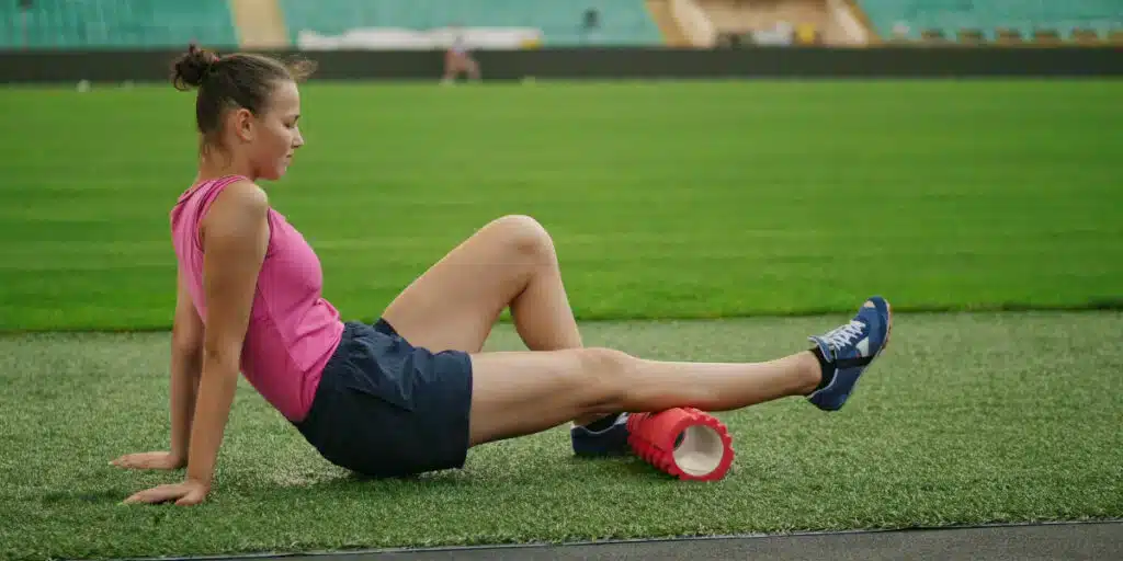 A woman is sitting on the grass foam rolling her right leg.