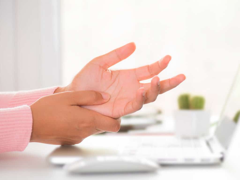 Eine rechte Hand umfasst das Handgelenk der linken, nach oben geöffneten Hand vor einem Laptop.