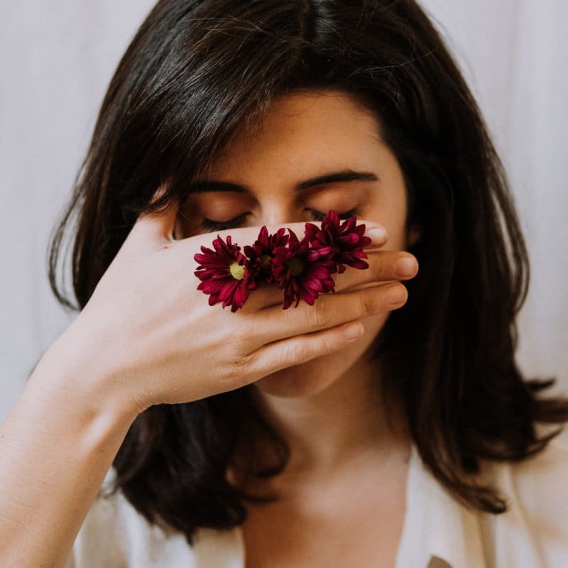 Eine schwarzhaarige Frau mit gesenktem Blick hält ihre Hand vor das Gesicht. Zwischen den Fingern stecken drei lilafarbene Blumen.