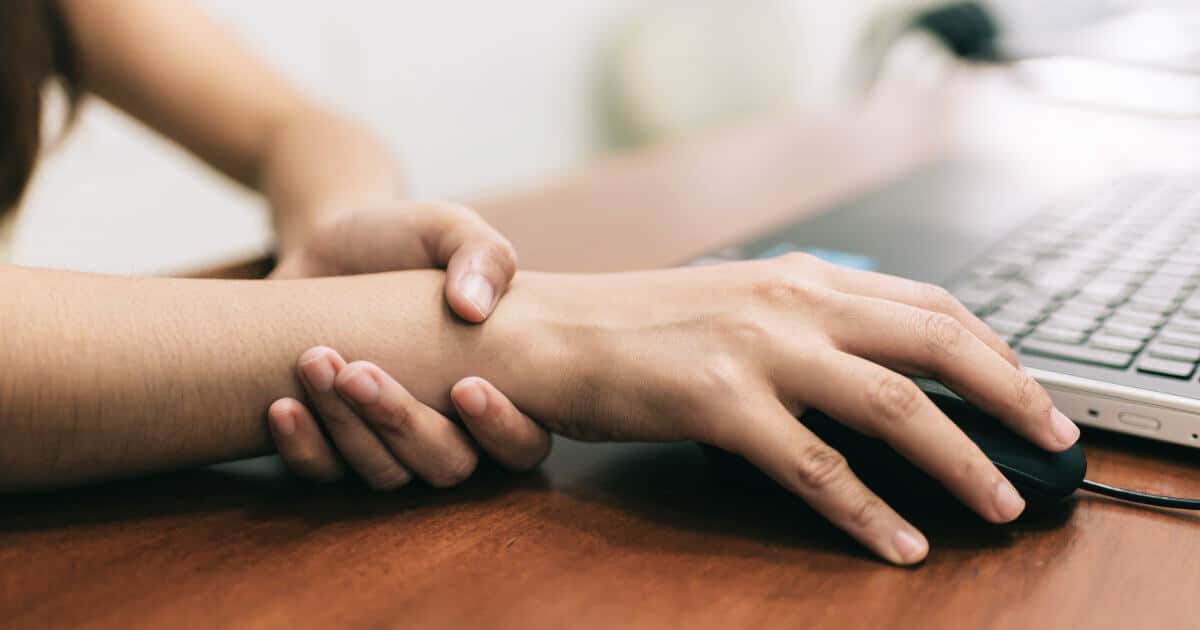Frau umgreift mit einer Hand das Handgelenk der anderen Hand. Mit dieser bedient sie eine Maus.