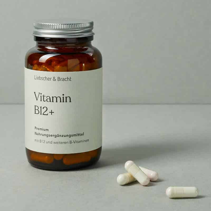 Eine kleine, dunkle Glasflasche steht vor einem dunkelgrünen Hintergrund. Sie ist mit Vitamin B12plus beschriftet. Daneben liegen vier Tabletten in heller Farbe.