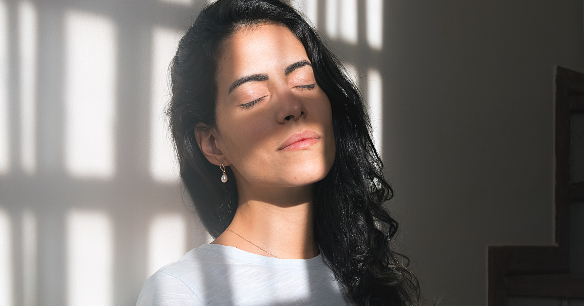 Eine schwarzhaarige Frau steht hinter einem Fenster an eine Wand gelehnt. Sie genießt die hereinfallenden Sonnenstrahlen und hat die Augen geschlossen, während sich auf ihrem Gesicht das schattige Gitter der Fenster abzeichnet.