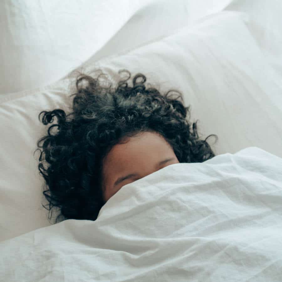Eine Frau liegt im Bett. Nur ein Teil ihres Kopfes, Haare und bis zum Ansatz ihrer Nase schauen unter der weißen Bettdecke hervor.