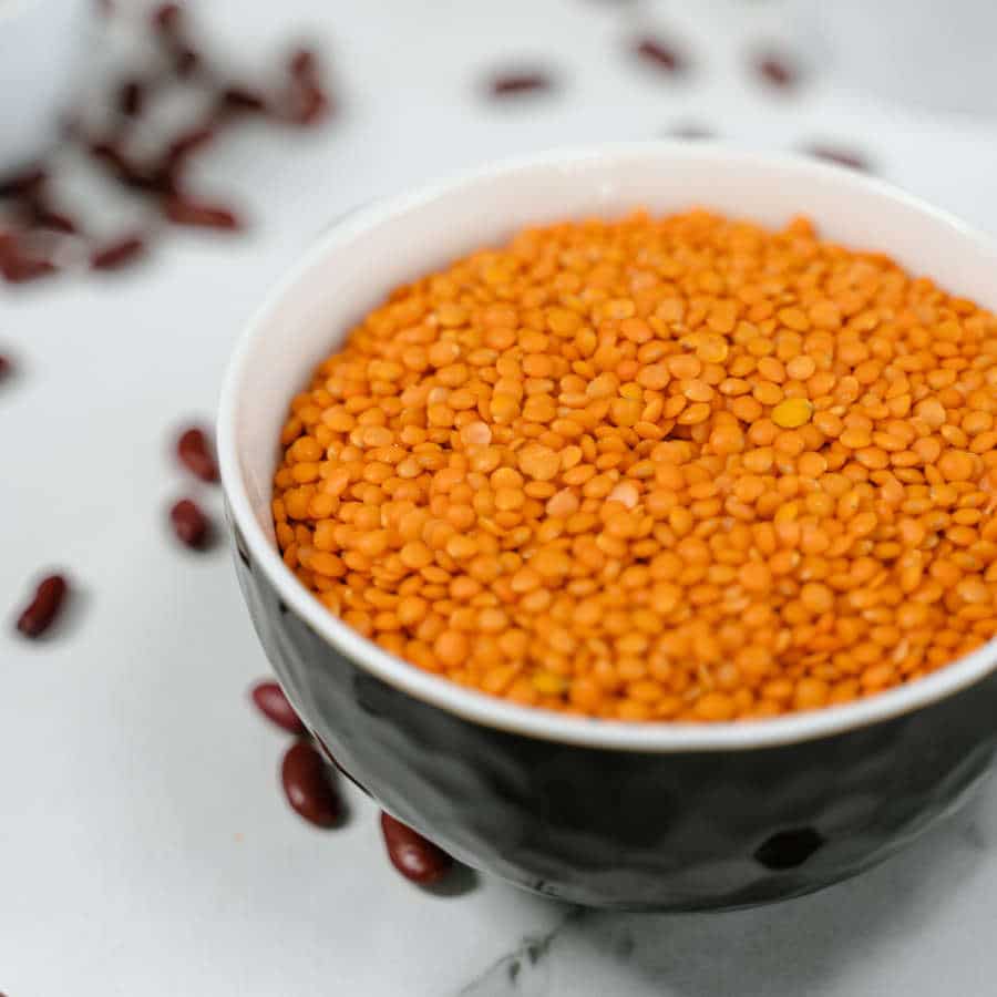 Rote Linsen sind in einer schwarzen Schale. Drum herum liegen rote Bohnen auf einem Tisch verteilt.