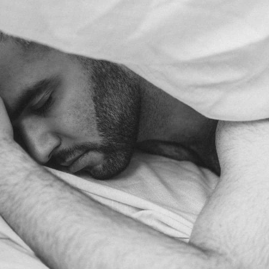 Ein Mann liegt unter einer weißen Decke und schläft auf dem Bauch. Zu sehen ist nur sein halber Kopf, den er auf dem Bett abgelegt hat und sein linker Arm, der angewinkelt neben seinem Kopf liegt.
