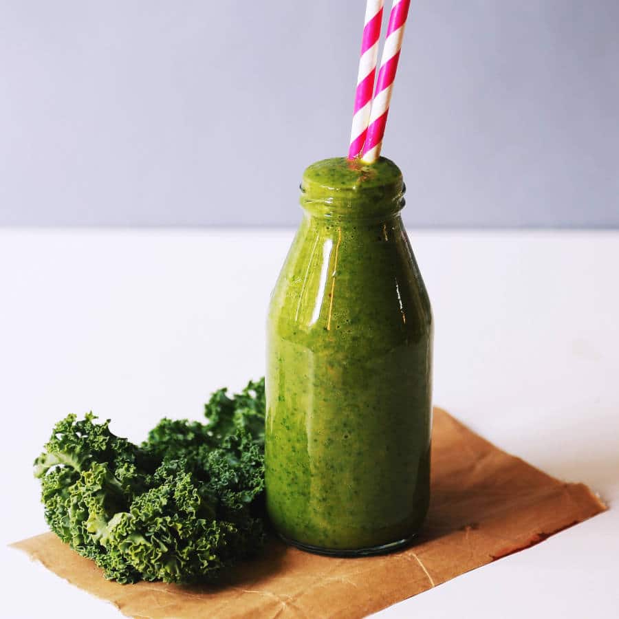 Ein grüner Smoothie mit Grünkohl ist in einer Glasflasche auf einem Tisch. Aus der Glasflasche ragen zwei pink-weiße Strohalme