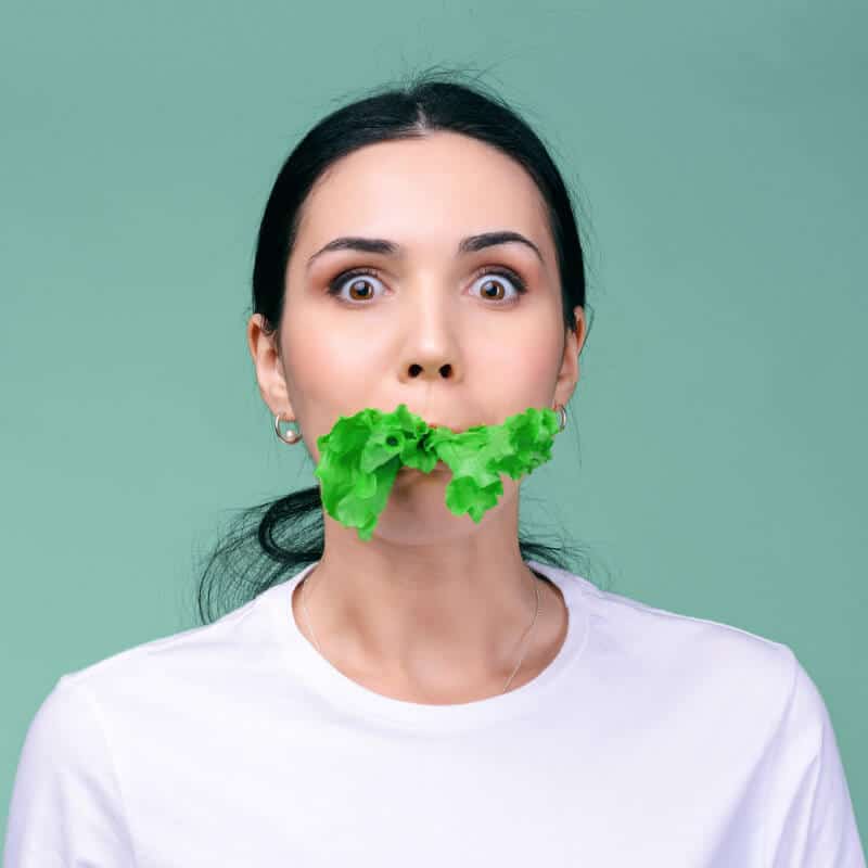 Eine junge Frau hat ein grünes Salatblatt im Mund und schaut erschrocken in die Kamera