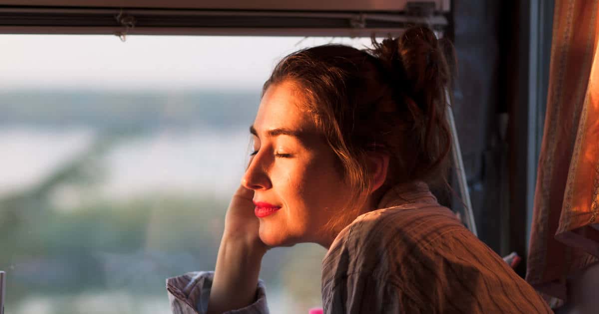 Junge Frau sitzt am Fenster eines Zuges. Sie hat die Augen geschlossen und genießt das Sonnenlicht, das ihr ins Gesicht scheint