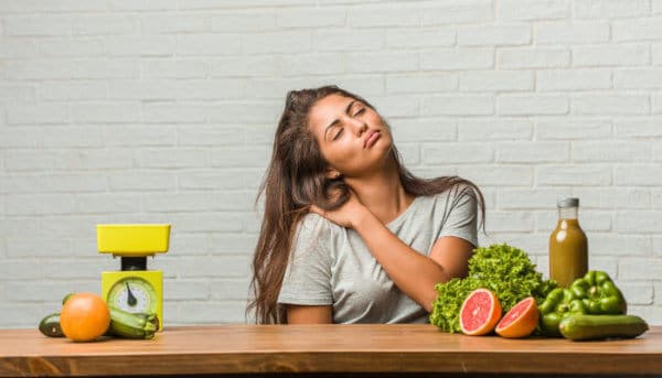 Eine Frau sitzt mit geschlossenen Augen an einem Tisch und berührt mit einer Hand ihre rechte Schulter. Auf dem Tisch liegen Paprika, Grapefruit, Zucchini, Orangen, Saft und eine Küchenwaage verteilt.