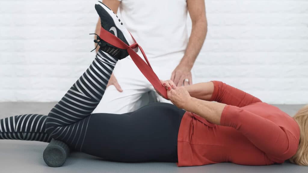 Eine Frau liegt auf dem Bauch. Ihr linkes Bein hat sie angewinkelt, unter dem Knie liegt die Medi-Rolle. Die rote Übungs-Schlaufe hat sie um ihren Fuß gefädelt. So kann sie sich das Bein näher zum Gesäß ziehen.