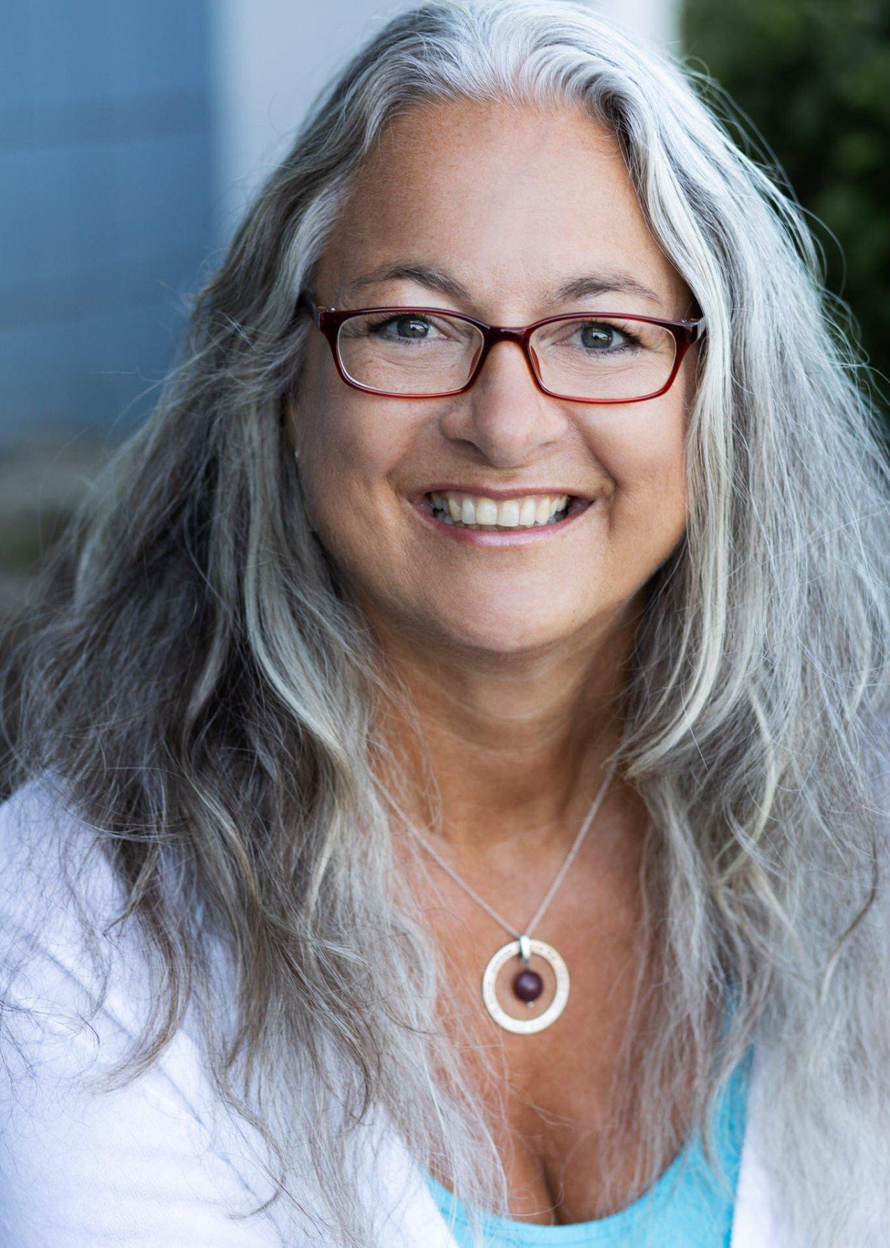 Birgit Fuchsenthaler, Qualitätspartnerin aus Neckargemünd, steht vor einem weißen Hintergrund. Sie hat lange graue Haare und trägt neben einem weißen Liebscher & Bracht T-Shirt, eine bunte runde Brille.