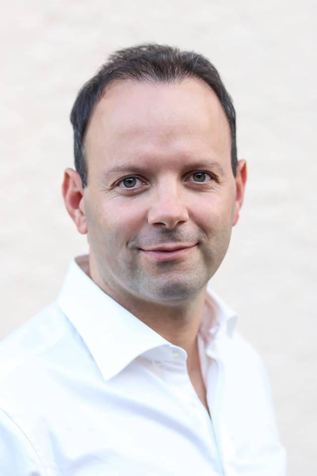 Christoph Schedler ist in Garmisch-Partenkirchen Physiotherapeut und steht vor einer hellen Wand. Er hat kurze dunkle Haare und trägt ein weißes Hemd.