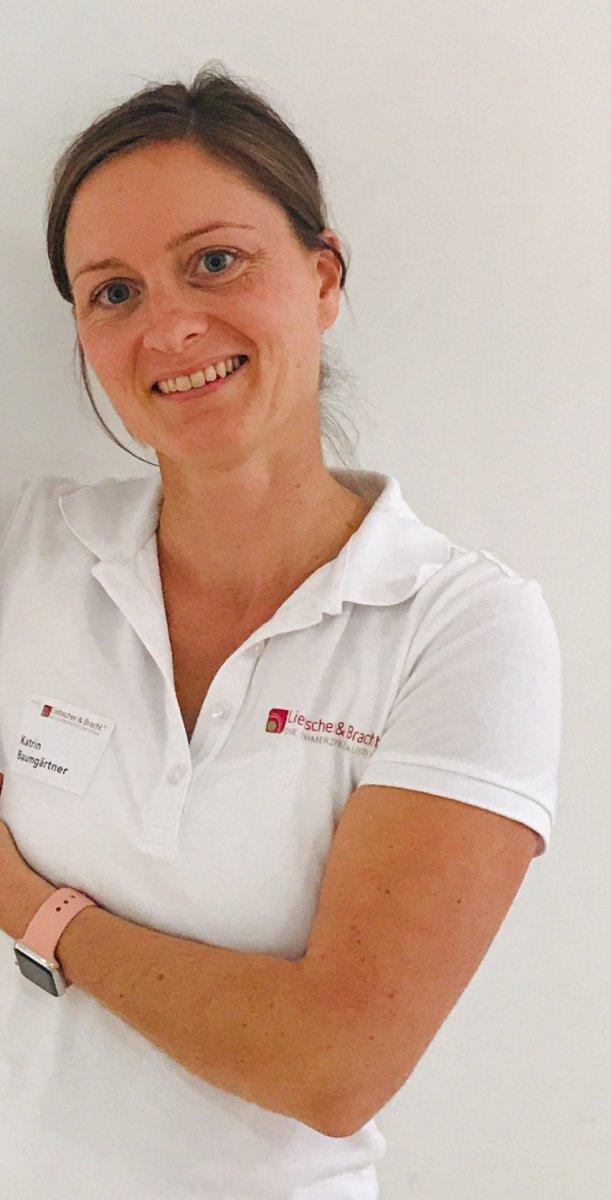 Die aus Bad Schönborn stammende Katrin Baumgärtner steht vor einem weißen Hintergrund. Sie hat ihre braunen Haare zu einem Zopf gebunden und trägt ein weißes Liebscher & Bracht T-Shirt. Sie lacht in die Kamera und hält einen Arm vor den Brustkorb.