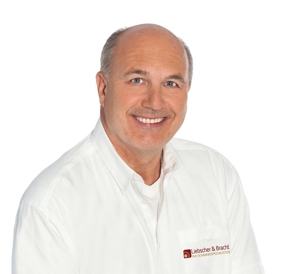 Dr. Markus Hundhammer ist Schmerztherapeut aus Grünwald. Er lächelt sympathisch, trägt ein weißes Liebscher & Bracht Hemd und hat eine Halbglatze mit grauen Haaren.
