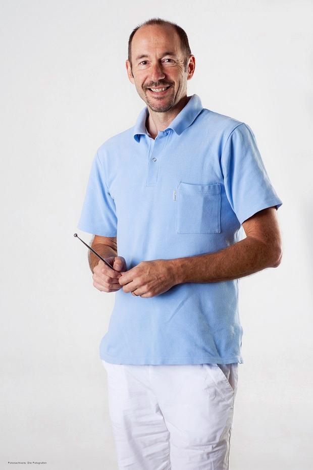 Auf dem Foto steht der Schmerztherapeut Peter Herndler vor einem weißen Hintergrund. Er hat kurze braune Haare und eine Halbglatze sowie einen Dreitagebart. Er trägt ein hellblaues Polo T-Shirt und eine weiße Hose.