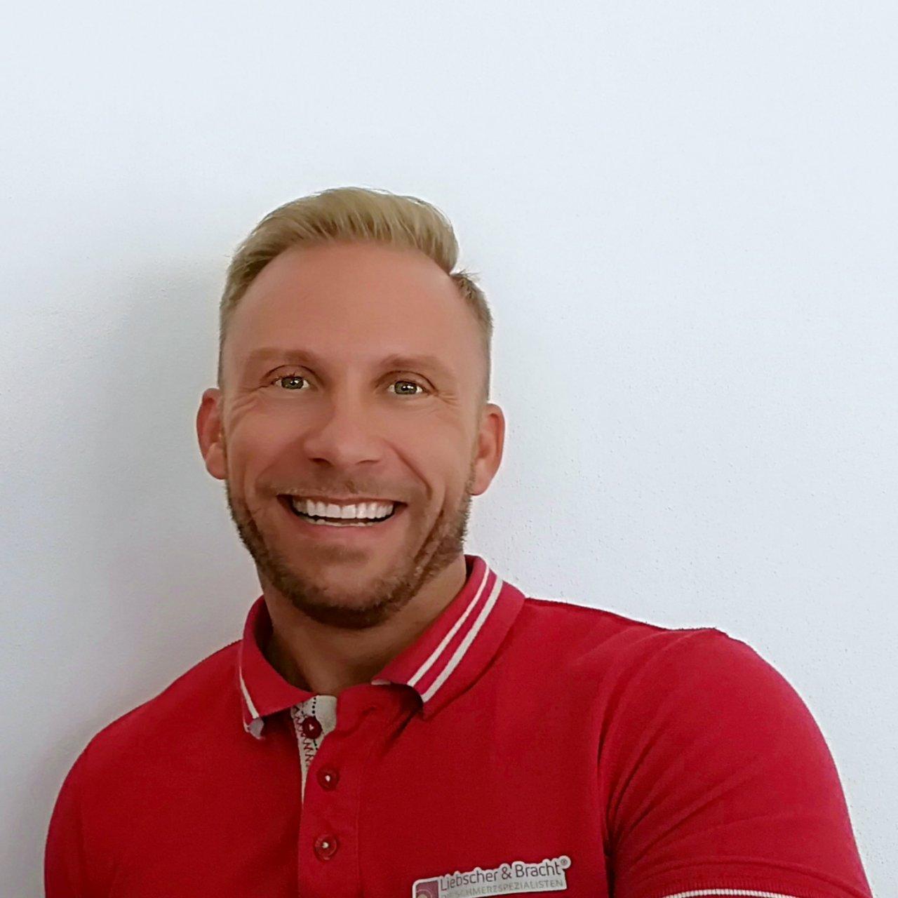 Das Foto zeigt den Physiotherapeuten Daniel Nenz. Er hat kurze blonde Haare und trägt ein weißes Polo T-Shirt.