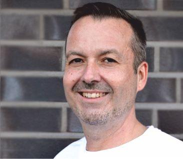 Auf dem Foto steht Markus Zach-Landvogt, der in Bad Zwischenahn praktiziert. Er steht vor einer dunklen Backsteinwand. Er hat dunkle Haare und einen Dreitagebart.