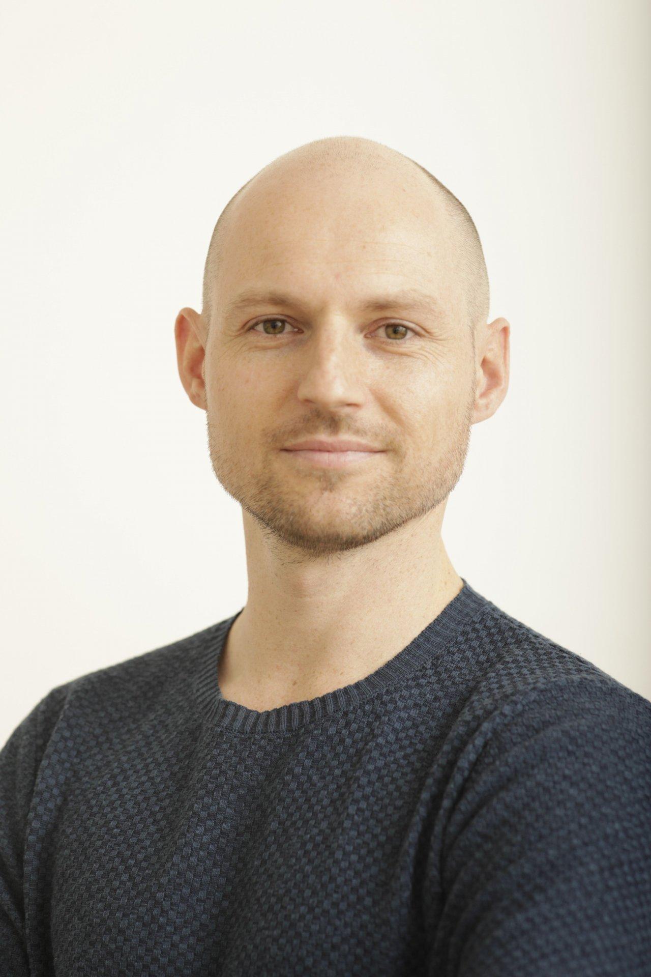 Der Diplom-Physiotherapeut aus Zürich Paul Hörter steht vor einem weißen Hintergrund. Man sieht ihn vom Oberkörper aufwärts. Er hat kurze blonde Haare und trägt ein Liebscher & Bracht Polo T-Shirt.