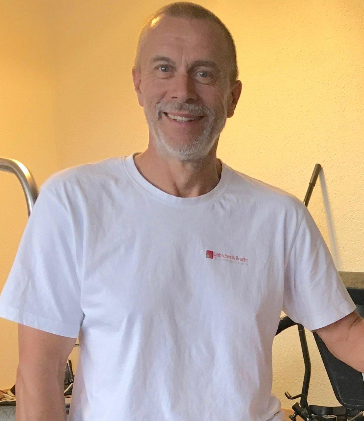 Physiotherapeut aus Wabern, Matthias Goering trägt ein weißes Liebscher & Bracht T-Shirt und steht vor einem gelben Hintergrund mit Fitnessgeräten.