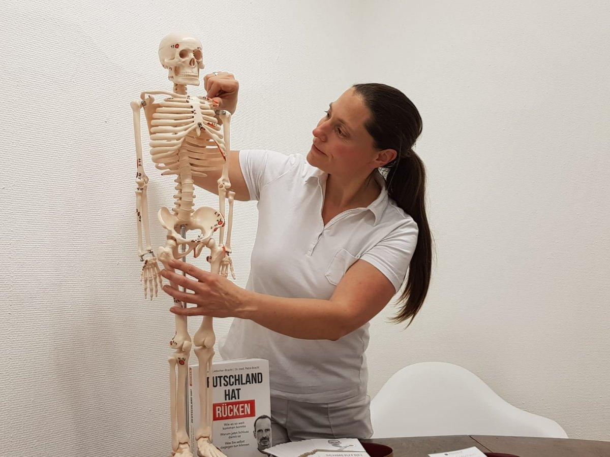 Das Foto zeigt Martha Holzhausen, wie sie an einem Skelett etwas erklärt. Sie hat lange schwarze Haare, die sie als Zopf trägt. Dazu hat sie ein weißes Polo T-Shirt an. Neben dem Modellskelett steht das Buch "Deutschland hat Rücken".