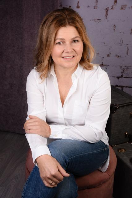 Das schwarz-weiß Foto zeigt Renate Schrittenlocher aus 82152 Planegg wie sie lächelnd in die Kamera schaut. Sie hat Ihre Arme auf einem Sessel aufgelehnt und auf einen Arm stützt sie ihren Kopf. Sie hat schulterlange braune Haare und trägt eine helle Blus