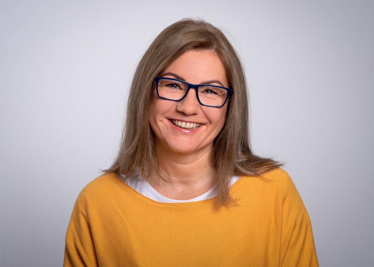 Auf dem Foto ist Aleksandra Riepe-Darsznik vor einem hellgrauen Hintergrund zu sehen. Sie hat hellbraune - graue schulterlange Harare und trägt eine dunkle eckige Brille. Sie trägt ein weißes T-Shirt und einen senfgelben Pullover und lächelt freundlich in