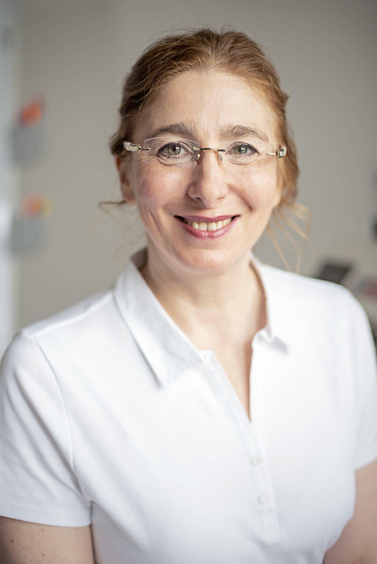 Auf dem Foto lacht Aleksandra Traub in die Kamera. Sie trägt ihre braunen Haare als Zopf und trägt eine randlose Brille. Weiterhin trägt sie ein weißes Polo T-Shirt.