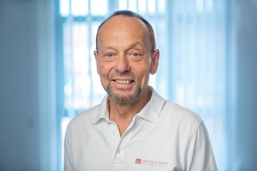 Auf dem Foto steht Andreas Enders aus Dieburg vor einem hellen Fenster. Er trägt ein weißes Liebscher & Bracht Polo T-Shirt und hat dunkle kurze Haare, sowie einen grauen Bart.