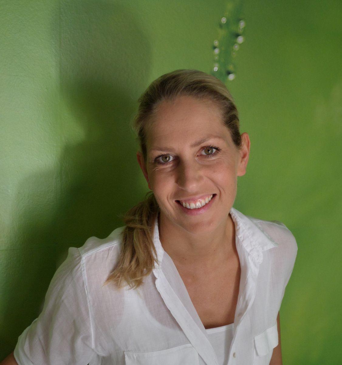 Auf dem Foto lehnt die Schmerz- und Bewegungstherapeutin aus 7053 Hornstein an einer grünen Wand. Katja Mössner lacht in die Kamera und trägt ihre langen blonden Haare zum Zopf, der auf der Schulter abgelegt ist. Sie trägt eine weiße Bluse.