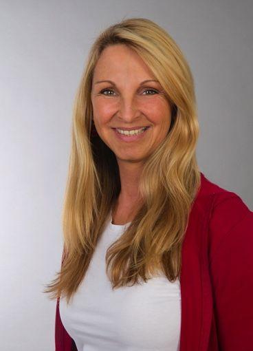 Sabine Hoeher aus Berlin steht vor einem hellgrauen Hintergrund. Sie trägt weiße Kleidung und eine rote Strickjacke. Die Heilpraktikerin und Qualitätspartnerin lächelt freundlich und hat lange blonde Haare.