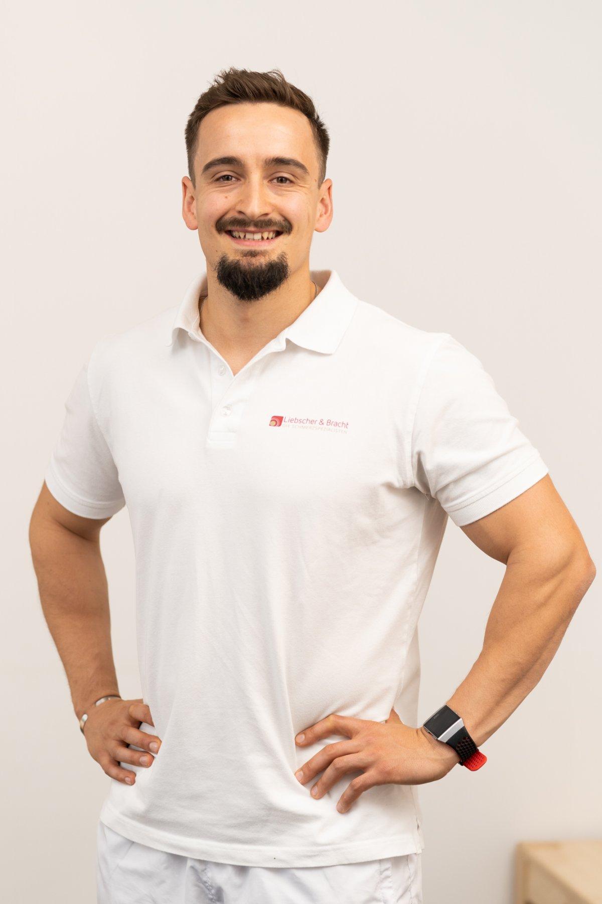 Auf dem Foto steht der sympathische, sportliche Simon Bösch vor einem weißen Hintergrund. Er trägt weiße Kleidung und ein Liebscher & Bracht T-Shirt und hat kurze braune Haare und einen Bart.