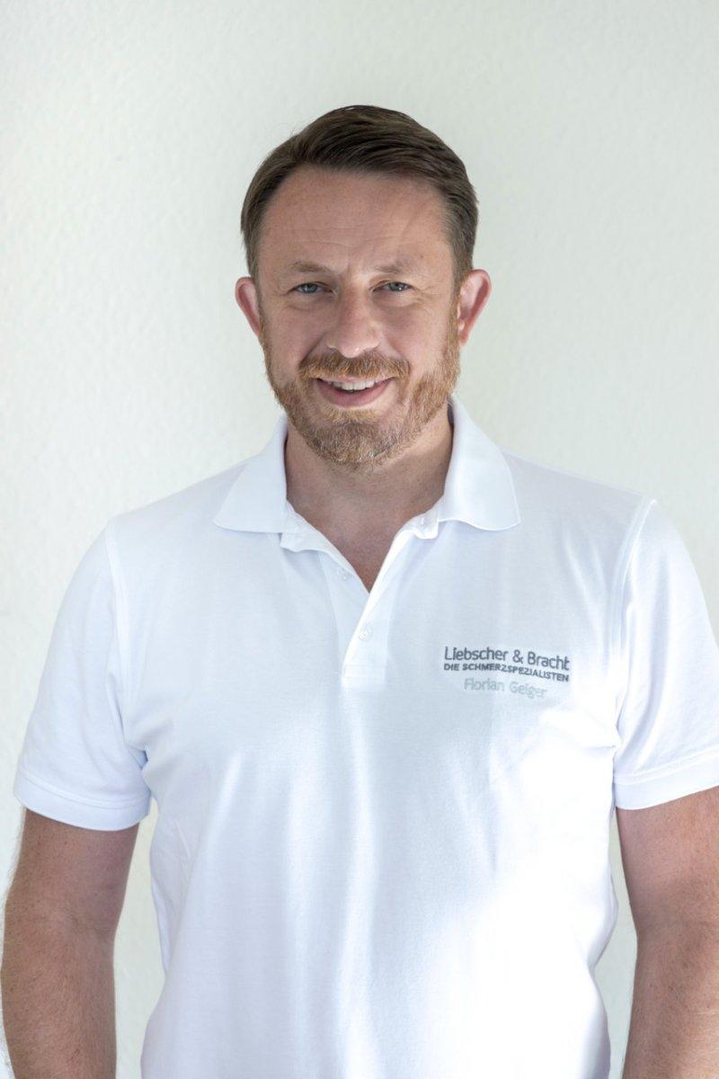 Florian Geiger, Schmerzspezialist und Pysiotherapeut aus Schäbisch Gmünd, steht vor einem weißen Hintergrund. Er hat braunes Haar und einen Vollbart. Zudem trägt er ein weißes Liebscher & Bracht Polo T-Shirt.