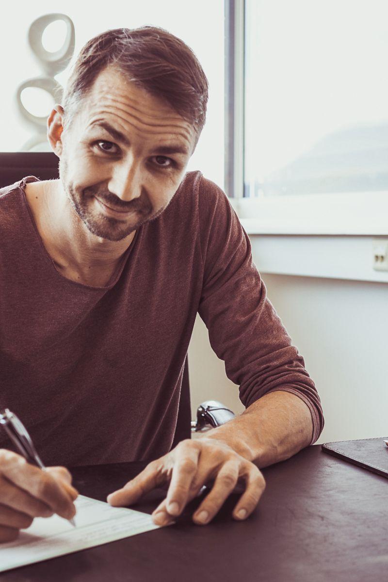 Auf dem Foto ist Herbert Scheipl aus Elixhausen zu sehen. Er sitzt am Schreibtisch und unterschreibt gerade ein Dokument, dabei schaut er lächelnd in die Kamera. Er hat kurze braune Haare und einen Dreitagebart und trägt ein braunes Shirt.