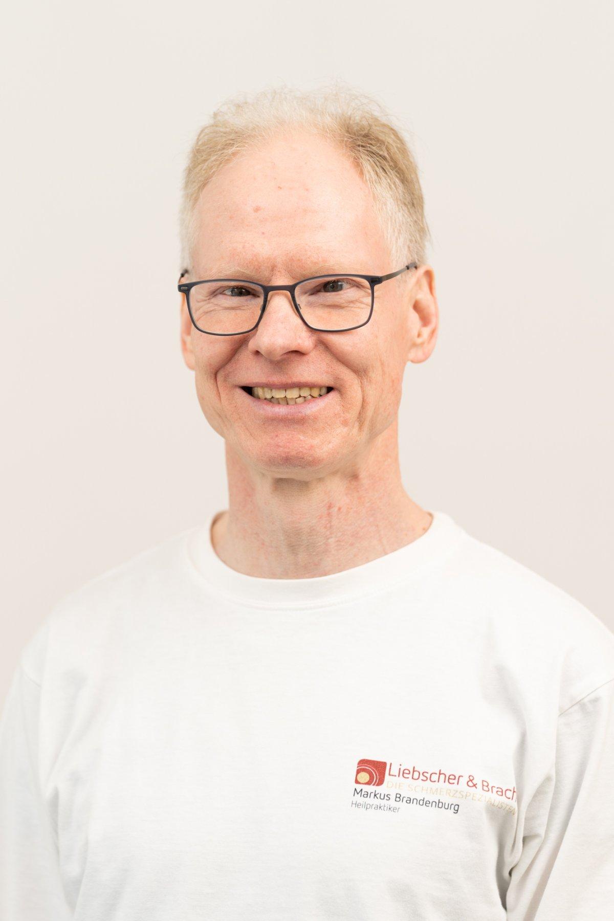 Der Heilpraktiker aus Berlin Steglitz Markus Brandenburg steht vor einem weißen Hintergrund. Er trägt ein Liebscher & Bracht T-Shirt, eine schwarze eckige Brille und hat kurze blond-graue Haare.
