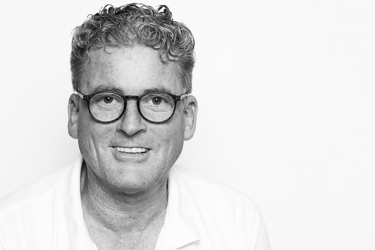 Das Foto zeigt Jürgen R. Hahn aus Reinbek. Der L& B Schmerzspezialist trägt ein weißes T-Shirt und längere Braun-graue Locken, die er nach hinten gebunden hat.