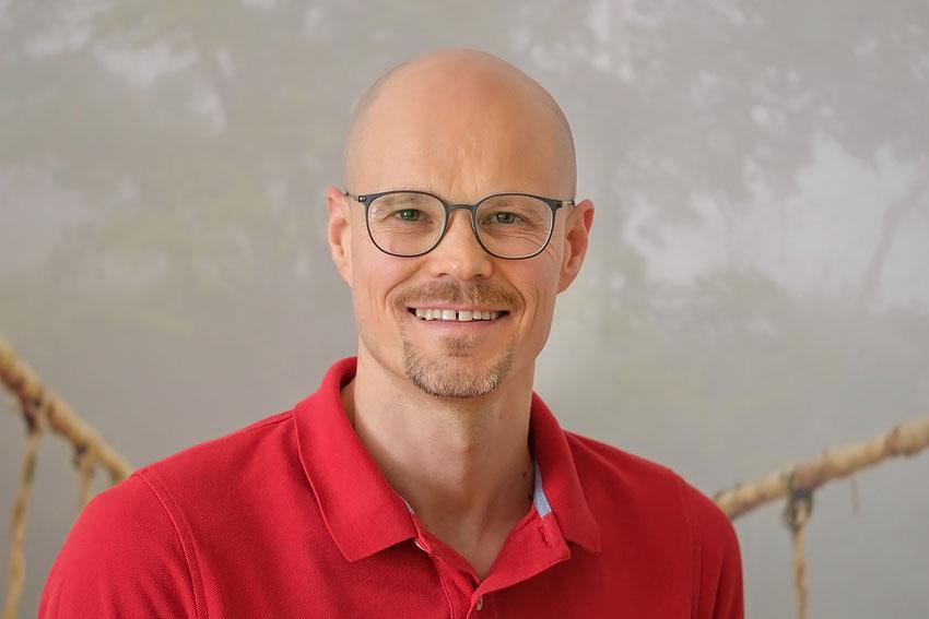 Das Foto zeigt den Heilpraktiker für Physiotherapie Sergej Rempel in einem Behandlungsraum. Er trägt eine schwarze randlose Brille und ein weißes Polo T-Shirt mit schwarzen Rändern.