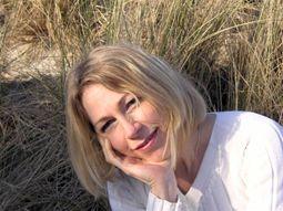 Myriam Ott-Jacquet sieht man vor dem Hintergrund einer Düne mit Dünengras sitzen. Sie hat schulterlange blonde Haare und lehnt ihren Kopf auf eine Hand. Sie lächelt in die Kamera und trägt eine weiße Bluse.