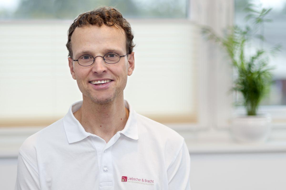 Der Ergotherapeut aus Hanstedt in der Nordheide sitzt vor einem lichtdurchfluteten Fenster. Martin Brandt trägt ein weißes Liebscher & Bracht Polo T-Shirt, hat braune kurze lockige Haare und eine schwarze runde Brille.