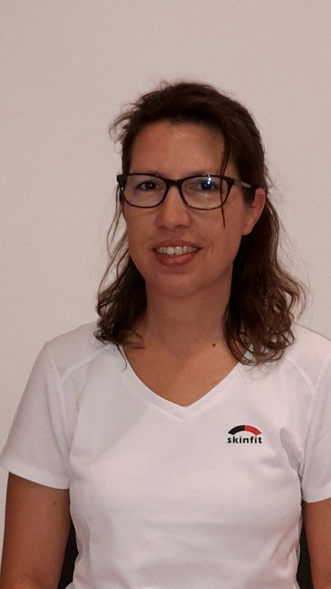 Das Foto zeigt Barbara Krähenbühl vor einem weißen Hintergrund. Sie hat schulterlange braune Haare, die sich leicht wellen und trägt eine eckige dunkle Brille sowie ein weißes T-Shirt mit Aufschrift.