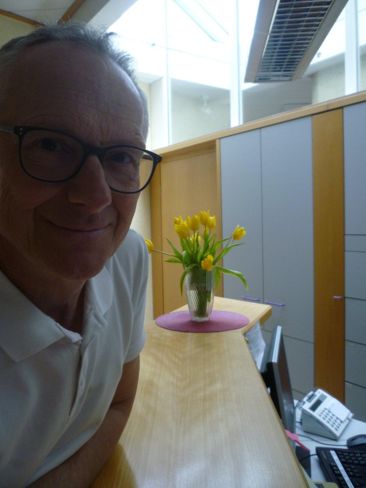 Auf dem Foto lehnt Dr. Thomas Werner aus Bad Königshofen lehnt an einer Theke. Im Hintergrund ist ein Blumenstrauß und ein Schrank zu sehen. Dr. Werner hat kurze Haare und trägt eine schwarze eckige Brille und ein weißes Polo T-Shirt.